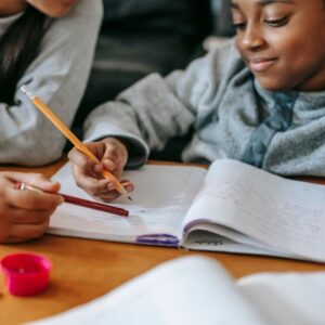 kid writes in notebook with pencil with instructor beside them 