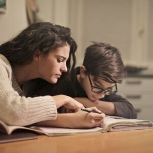 kid wearing glasses leans over notebook on table with woman leaning in beside 