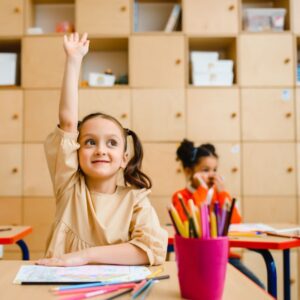 girl with pigtails raises hand sitting at desk 